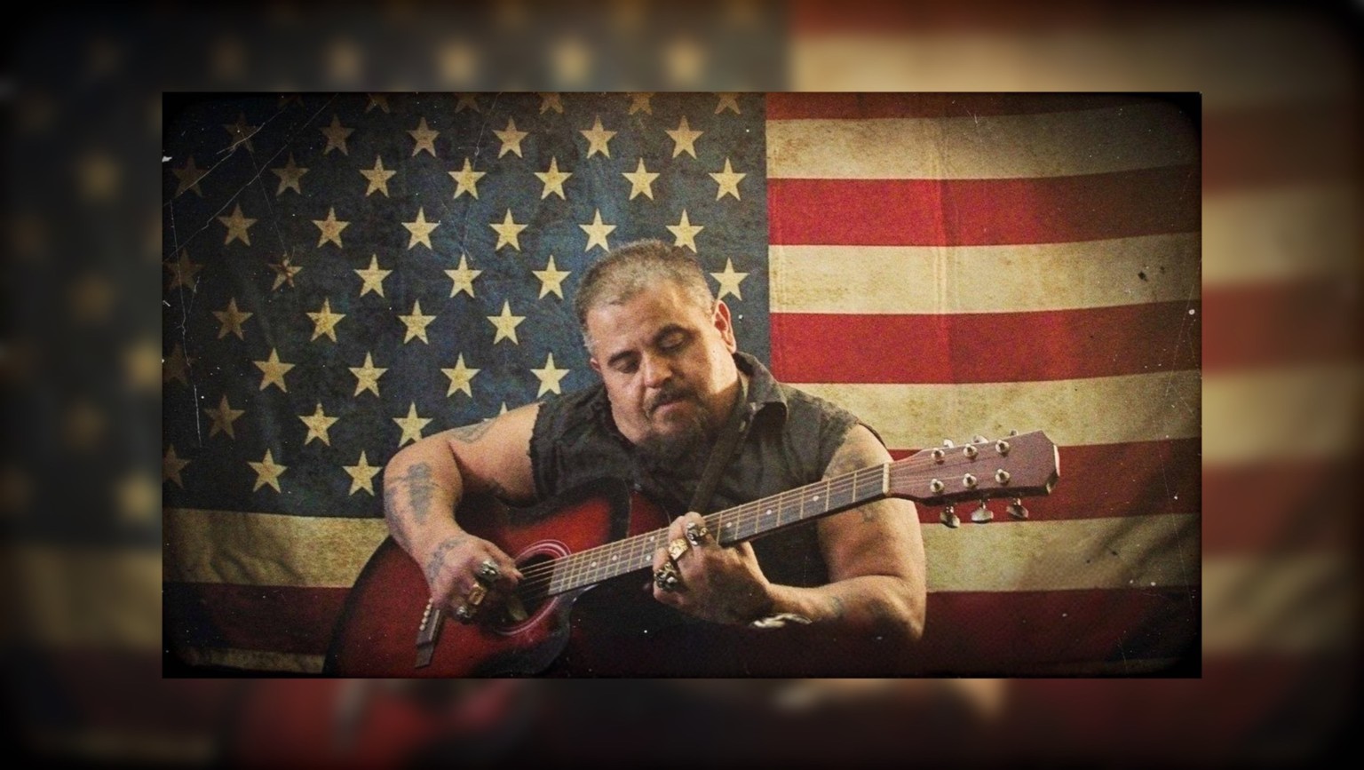 Preacher Joe playing acoustic guitar in front of an American flag