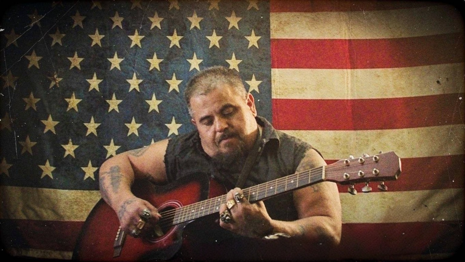Preacher Joe playing acoustic guitar in front of an American flag